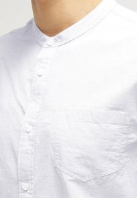 Hombre vestido con una camisa blanca de cuello mao, con botones en la parte delantera y un bolsillo en el pecho.
