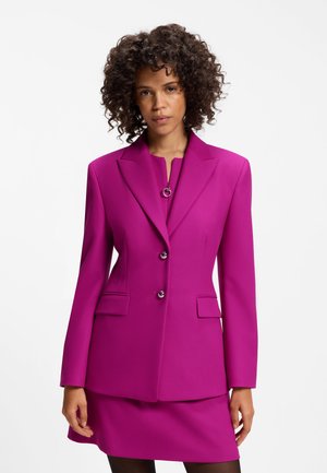 Kvinde med krøllet hår iført en tætsiddende, lys magenta blazer med to knapper og matchende nederdel mod en ensfarvet hvid baggrund.