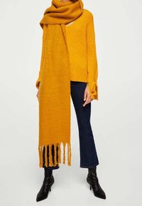 Kobieta w musztardowym, oversize'owym szaliku i pasującym swetrze, ciemnych, krótkich dżinsach oraz czarnych, skórzanych botkach na obcasie.