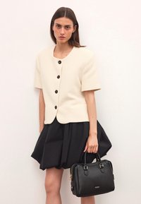 Kermakuvioinen, lyhythihainen takki, jossa on pyöreä kaula-aukko ja mustat napit; yhdistettynä mustaan, laskostettuun hameeseen ja pieneen mustaan käsilaukkuun.