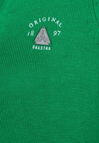 Zelená pletená tkanina s trojúhelníkovou šedou nášivkou, nad kterou je bílý nápis "ORIGINAL 1897 GAASTRA". Texturovaný povrch.