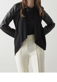 Veste noire drapée avec des manches en cuir, présentant une texture lisse et un design à l'avant ouvert, portée sur un top noir et un pantalon blanc.