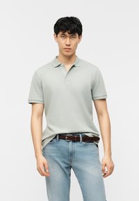 Νεαρός άνδρας φοράει ανοιχτό πράσινο πόλο πουκάμισο, μαζεμένο μέσα σε ανοιχτόχρωμα μπλε τζιν, με καφέ πλεκτή ζώνη, στέκεται μπροστά σε ένα απλό λευκό φόντο.