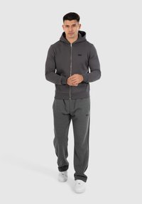 Hoodie zippé gris avec un logo discret, associé à un pantalon de survêtement gris foncé avec des bandes latérales, complété par des baskets blanches.
