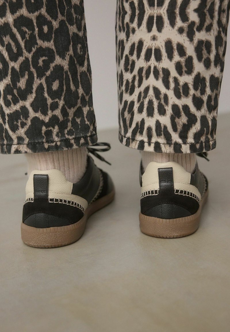 Schwarze und cremefarbene Sneaker mit einer strukturierten Oberfläche. Akzentuiert mit Nähten und einer braunen Gummisohle. Getragen mit gemusterten Leopardenprint-Hosen.