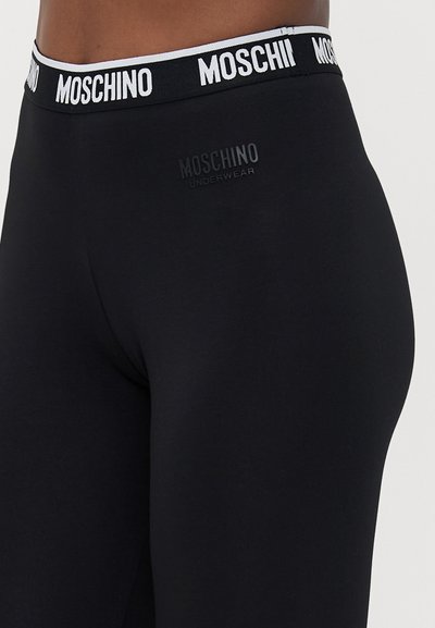 Musta, kehaümberkättev pesu Moschino bränditud vöökoht ja logo vasakul puusal, kantud inimese madalal kehapiirkonnas.