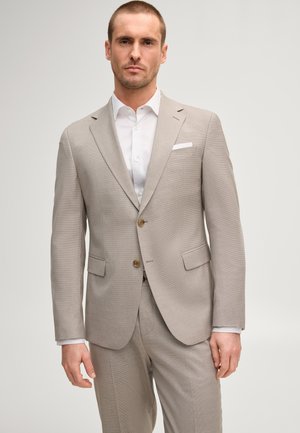 Uomo che indossa un abito beige con camicia bianca e fazzoletto da taschino, in piedi davanti a uno sfondo chiaro e semplice.