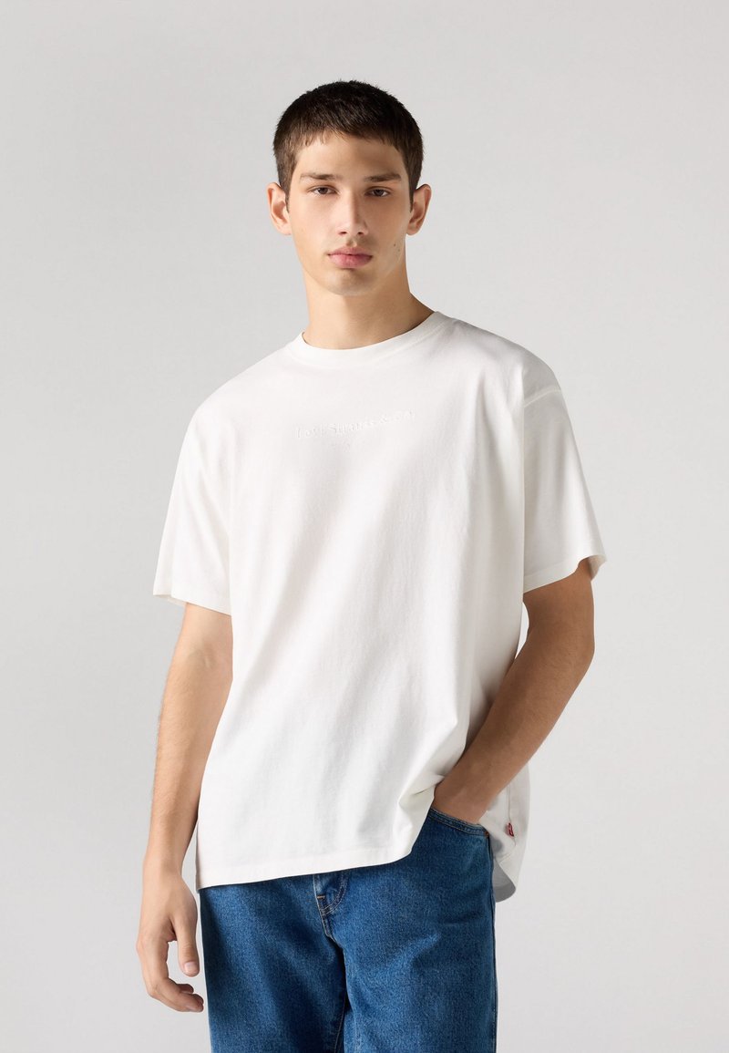 Weißes Baumwoll-T-Shirt mit kurzen Ärmeln, rundem Ausschnitt und geprägtem Logodetail. Kombiniert mit blauen Jeans. Einfaches und minimalistisches Design.