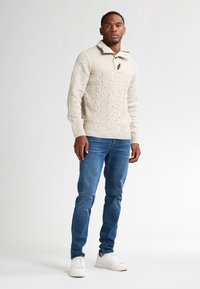 Neulepaita vaaleanbeigessä, jossa on teksturoitu palmikkokuvio, neljännes-vetoketjukaulus ja puunappi; yhdistettynä sinisiin farkkuihin ja valkoisiin tennareihin.