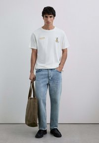 Bărbat îmbrăcat cu un tricou alb cu grafică, blugi de un albastru deschis și pantofi negri, ținând o geantă de umăr maro într-o mână, cu o mână în buzunar, pe un fundal simplu.
