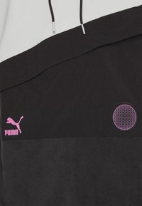 Gros plan d'un sweat à capuche noir et blanc avec un logo Puma rose et un patch violet à motifs circulaires à l’avant.