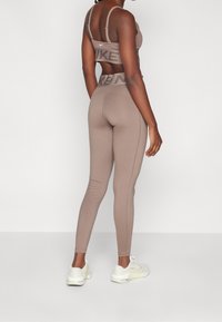 Hnědé sportovní legíny s středně vysokým pasem, zdobené texturovaným logem Nike; doplněné odpovídajícím sportovním podprsenkou a bílými teniskami.