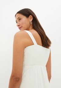 Abito bianco con corpino arricciato, spalline larghe e tessuto fluente. I capelli sono lisci e castani, acconciati in modo morbido. Orecchini a cerchio in argento visibili.