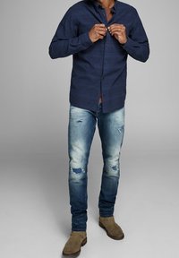 Marinrandig skjorta med knappar, ljusblå slitna jeans med rivna lappar och bruna mockaskor över anklarna. Avslappnad passform och strukturerad tyg.