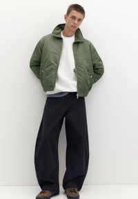 Jeune homme portant une veste vert olive, un sweat-shirt blanc, un pantalon large noir et des chaussures marron, debout devant un fond blanc uni.