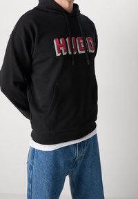Felpa nera con cappuccio caratterizzata da una tasca a marsupio frontale, logo "HUGO" in rosso e verde, polsini e orlo ribattuti, abbinata a jeans blu.