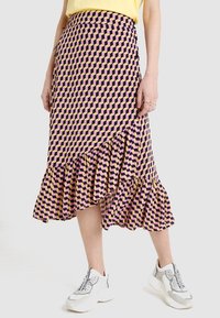 Gemusterter Midirock in lila und cremefarbenem geometrischem Design, mit einem Rüschensaum und einer taillierten Passform, kombiniert mit weißen Sneakern.