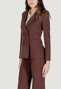 Hnědý dvouřadý blazer s klopami, dvěma předními kapsami a odpovídajícími upravenými kalhotami. Hladká tkanina s přiléhavým střihem.