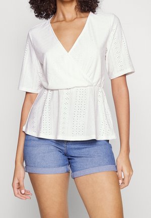 Kobieta ubrana w biały, krótkim rękawem, ażurowy top na zakładkę i niebieskie dżinsowe szorty ze zwiniętym dołem, stojąca na jednolitym tle.