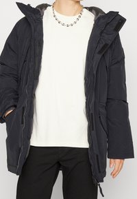 Personne portant une doudoune noire ouverte sur une chemise blanche, un pantalon noir et un collier chaîne argenté, debout avec une main dans la poche.