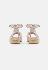 Espadrilles de color beige claro con suela de yute trenzado y lazos de tela en el tobillo, con un diseño de talón abierto y material superior suave y texturizado.