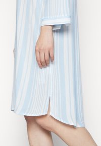 Vestido listrado azul claro e branco com um corte solto, apresentando uma abertura lateral, mangas longas e um tecido suave e texturizado.