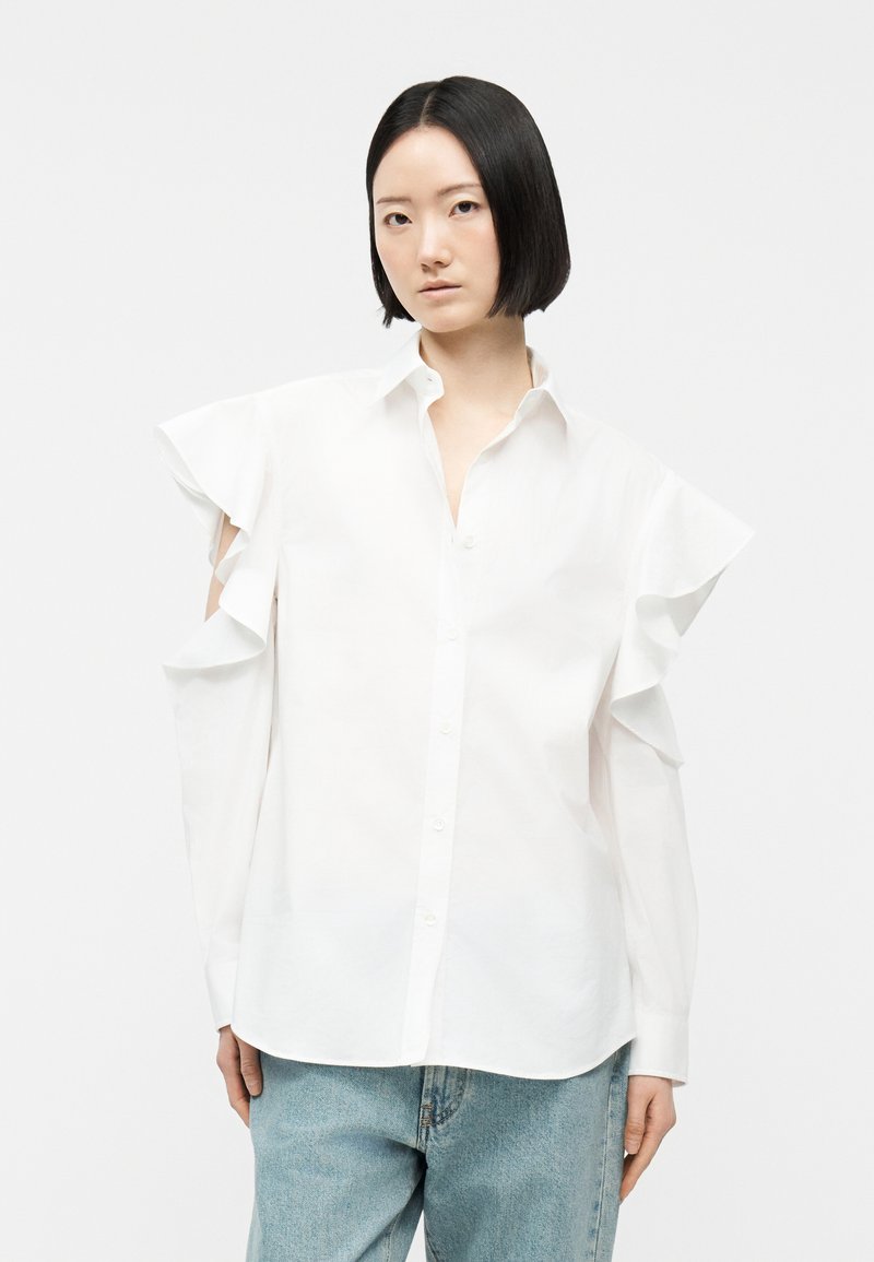 Tânără cu păr negru scurt, purtând o cămașă albă cu nasturi, cu detalii cu volane și umeri decupați, și blugi bleu deschis.