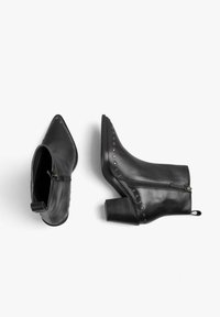 Botas de cuero negro hasta el tobillo con punta afilada, tachuelas decorativas plateadas a lo largo del costado y cierre de cremallera. Tacón grueso y texturizado.