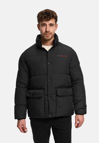 Jonge man met kort krullend haar, gekleed in een zwart donsjack met drukknopen en klepzakken, staand tegen een effen achtergrond.