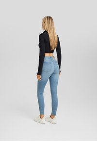 Top corto de canalé negro, jeans ajustados azul claro y zapatos blancos. Vista desde la parte trasera, destacando el ajuste de los jeans y el diseño del bolsillo trasero.