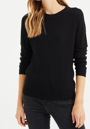 Woman wearing black textured long-sleeve sweater and black jeans, standing with her arms relaxed by her sides.