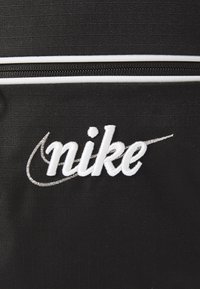 Črn nahrbtnik iz tkanine z izrazitim belim vezenim logotipom "nike" in belim trakom ob zadrgi. Tekstura se zdi odporna.