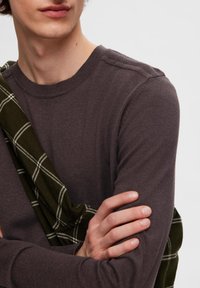Temno sivi dolgi pulover iz pletenine z rebrastim izrezom, nošen z zelenim karirastim šalom, ki je položen čez ramo. Gladka tekstura, prilegajoča oblika.