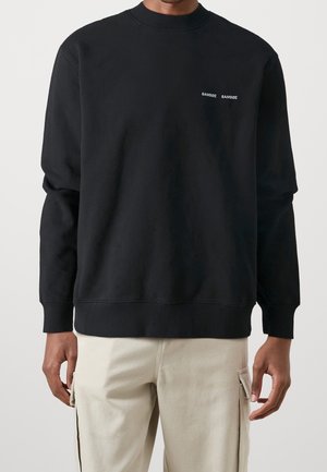 Mann trägt schwarzen Crewneck-Sweatshirt mit kleinem weißen "SAMSØE SAMSØE" Logo auf der Brust und hellbeigen Cargohosen.