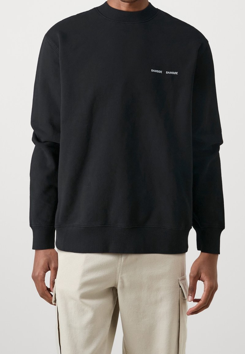 Homme portant un sweat-shirt noir col rond avec un petit logo blanc "SAMSØE SAMSØE" sur la poitrine et un pantalon cargo beige clair.