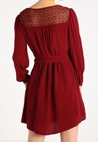 Bordo mekko, jossa on teksturoitu pitsiyläosa, pitkät hihat, kiristetty vyötärö ja laskeutuva helma. Sopivan sileä kangas, jossa on hillitty kiilto.