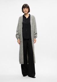 Cardigan largo de frente abierto en gris, hecho de un material de punto suave. Presenta mangas largas, dos bolsillos frontales y un corte relajado. Combinado con pantalones negros.