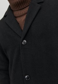 Cappotto di lana nera con una finitura texturizzata, revers a doppio petto e due bottoni neri. Sotto è indossato un dolcevita marrone a coste.