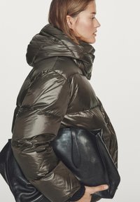 Olive green puffer jacket with a high collar, made of shiny material, and layered design. Black leather bag held under arm.