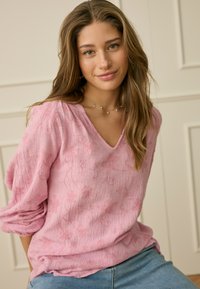Jeune femme aux longs cheveux bruns portant un chemisier rose texturé et un jean bleu clair, assise contre un mur beige à panneaux.