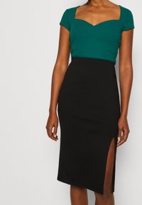 Zielony prążkowany top z kwadratowym dekoltem, połączony z czarną ołówkową spódnicą z bocznym rozcięciem dla dodatkowego efektu. Gładka faktura materiału.