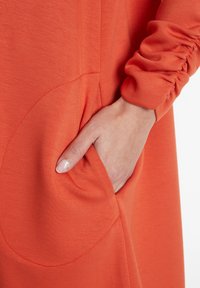 Orangene Kleid aus einem weichen Material. Auffällige Merkmale sind eine Tasche mit abgerundetem Rand und ein gerafftes Ärmeldetail.