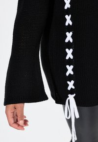 Schwarzer Strickpullover mit Glockenärmeln, der an der Seite mit weißen Schnürdetails versehen ist, ergänzt durch lose weiße Bänder am Saum.