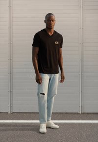 Μαύρο μπλουζάκι με κοντά μανίκια και V λαιμόκοψη με λογότυπο, ανοιχτό μπλε τζιν με σκισμένο γόνατο και λευκά αθλητικά παπούτσια. Στέκεται μπροστά σε γκρι φόντο.