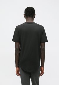 Jonge man met kort krullend haar, gekleed in een eenvoudige zwarte t-shirt met korte mouwen en donkergrijze broek, staand met zijn rug naar voren.