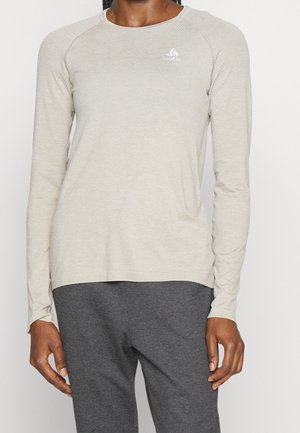 Osoba w beżowej, długorękawowej sportowej koszulce z małym logo na piersi oraz ciemnoszarych spodniach, stojąca na jednolitym tle.