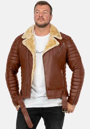 Mann trägt braune Lederjacke mit beigem Fellkragen, weißes Grafik-T-Shirt und schwarze Jeans, steht vor schlichter weißer Wand.