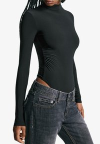 Kvinna som bär en figurnära svart l�ångärmad polotröja kombinerad med lösa jeans i mörk tvätt, poserande framför en enkel bakgrund.
