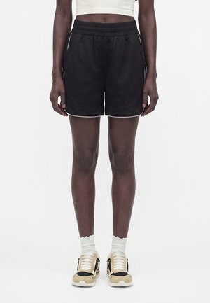 Άτομο που φοράει μαύρα αθλητικά σορτς από δίχτυ, λευκές κάλτσες αστραγάλου, μπεζ και μαύρα αθλητικά παπούτσια και λευκό κοντό τοπ, στέκεται μπροστά σε λευκό φόντο.