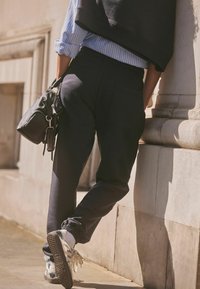 Marina joggers med elastisk midja och fållade benslut, i kombination med en ljusblå randig skjorta och en svart handväska, vilande mot en vägg.